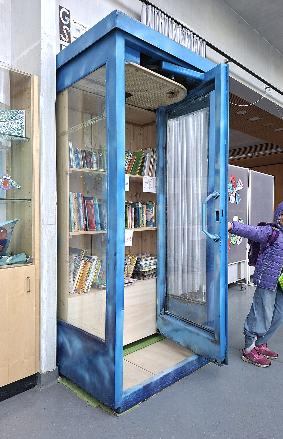 Bücherschrank In einer Aula steht eine blau angesprühte Telefonzelle. In der Telefonzelle sind Holzregale mit Kinderbüchern.